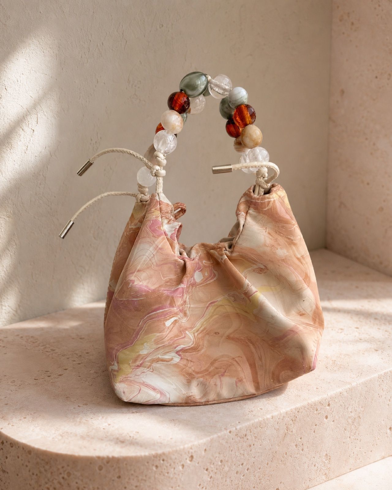 Peach Cloud soft marbled bag with multicolored bead handle on a warm stone plinth